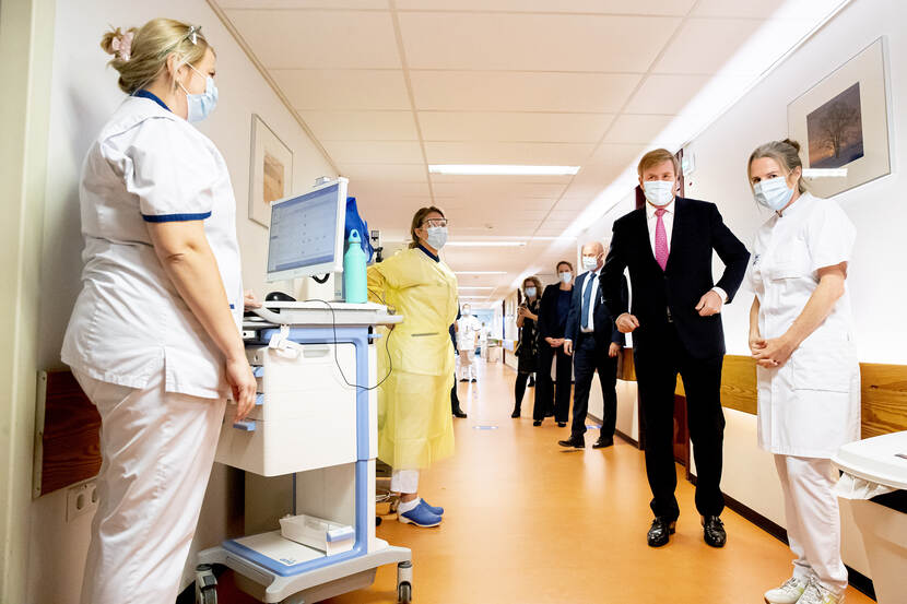 Koning in de gang van een ziekenhuis in gesprek met meerdere mensen. Zij dragen allen mondkapjes.