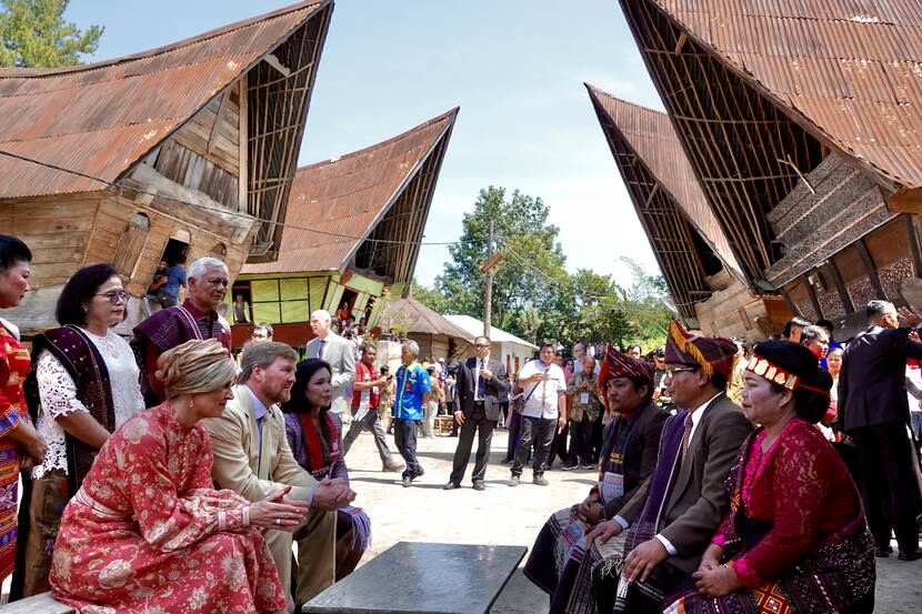 Koning Willem-Alexander en Koningin Máxima spreken met bewoners van een dorp op Sumatra. Op de achtergrond zijn vier traditionele Dajak-huizen te zien